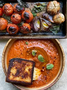 Overhead view of a baking sheet filled with blistered roma tomatoes, charred red onions, and roasted garlic cloves for roasted tomato soup.
